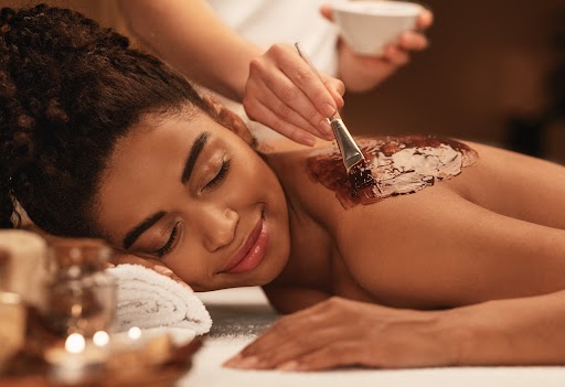 Young black woman receiving body scrub treatment at luxury spa, therapist applying exfoliating mask on female back, closeup